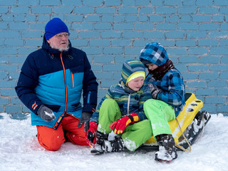 Fototapeta premium elderly man in bright winter clothes rides three boys in beautiful jackets on yellow sleds. Children love father. Winter games with snow. Kids enjoy spending time on vacation in village
