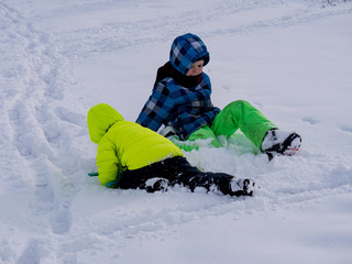 funny children in bright winter jackets and trousers walk in snowy field near village. boys lie on snow, jump, somersault, waving their hands depicting snow angels