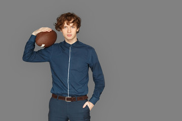 Sporty Lifestyle. Teenager boy standing isolated on grey with american football ball hand in pocket posing to camera confident