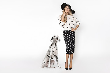 Attraktives, stylisches blondes Modell in schwarz-weißer Kleidung steht in der Nähe eines dalmatinischen Hundes. Schönes Model, gekleidet in einem schwarzen Kleid im Pinup-Stil mit weißen Tupfen, isoliert auf weißem Hintergrund © marcink3333