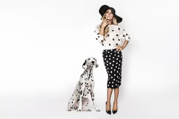 Attraktives, stylisches blondes Modell in schwarz-weißer Kleidung steht in der Nähe eines dalmatinischen Hundes. Schönes Model, gekleidet in einem schwarzen Kleid im Pinup-Stil mit weißen Tupfen, isoliert auf weißem Hintergrund © marcink3333
