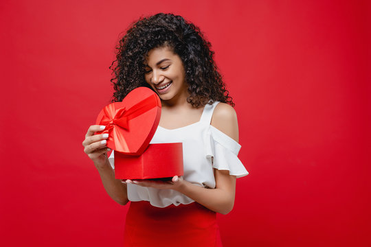 pretty cheerful black woman opening heart shaped valentine gift isolated on red