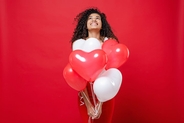 happy smiling black woman holding colorful heart shaped balloons isolated on red © Anastasia