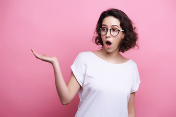 Good looking young Caucasian woman in round transparent eyewear, keeps hand raised, dressed in casual outfit, pretends holding something wonderful, isolated over pink background. Look there