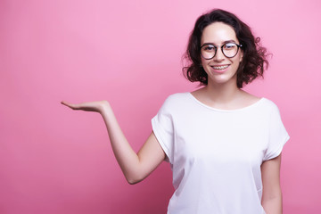 Good looking young Caucasian woman in round transparent eyewear, keeps hand raised, dressed in casual outfit, pretends holding something wonderful, isolated over pink background. Look there