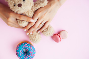 woman hands with manicure wearing wedding ring holdind teddy bear and macaroons on pink background