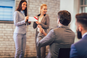 Front view of woman receiving award from businesswoman in front of business professionals applauding at business seminar in office building