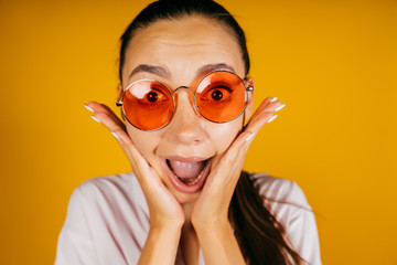 The emotion of amazement on the girl's face in pink glasses, her eyes are wide, her eyebrows are raised, her mouth is wide open, her palms hold her cheeks. background yellow