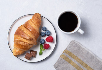 Fresh croissant on a white plate with blueberries and raspberries with a Cup of coffee on a gray background.