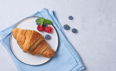 Fresh  croissant on a white plate and a blue napkin with berries  on a blue background.