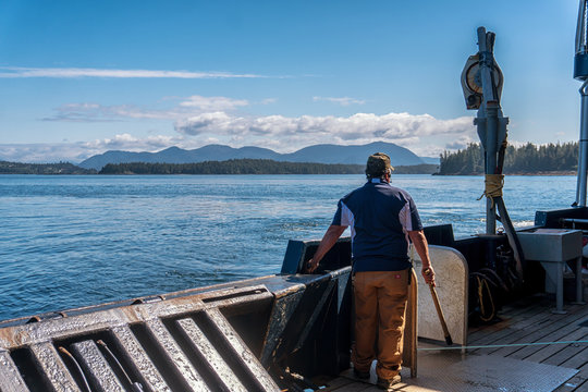 Single fisherman is standing on a commercial crab fishing boat and waiting to pull a pot. Special vessel in Bering sea for catching a king crab. Mountains and forest is on a background.