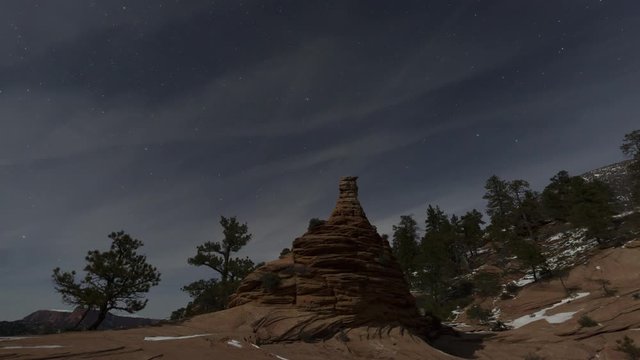 Winter stars slowly spin around the north star and thin clouds drift over the landscape of a sandstone hoodoo and pinyon trees with patches of snow here and there.