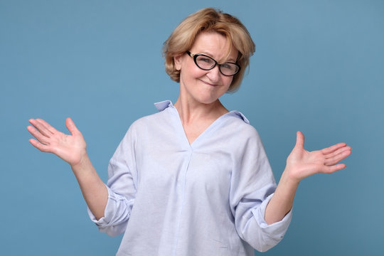 Senior mature caucasian woman in glasses and casual clothes shrugging her shoulders. Have no idea what to do in this situation. Studio shot