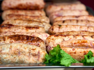Rows of freshly grilled chicken meat sausages on tray with fresh lettuce at street food festival, selective focus, close-up. Unhealthy oily barbeque street fast food for meat lovers concept