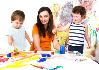 Woman with two babies painting with paints and brushes while sitting at childrens table, colorful background