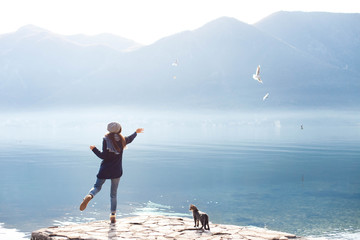 Young woman feeds seagulls and cat at winter sea beach. Amazing coastline scene with girl, animals, mountains, fog, blue water and morning light. Concept of freedom, travel, flying. Lifestyle moment.