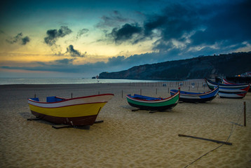 Portugal, twilight over Nazare beach , colored wooden boats, Traditional Portuguese fishing boats in Nazare on the coast, blue clouds on the sky, white sand of  beaches of Portugal