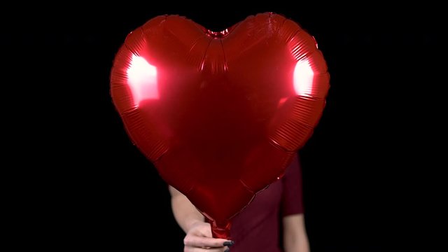 A young woman holds out a heart-shaped balloon towards the camera. A woman is standing with a helium ball on a black background. Valentine's Day is the day of all lovers.