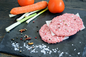 Fresh uncooked hamburgers with tomatoes, carrot, fresh green garlic and spices on the black kitchen board.