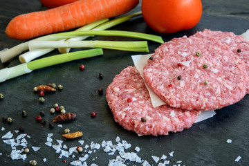 Uncooked fresh hamburgers with carrot, tomatoes, green garlic and spices on the black kitchen board prapared for cookin closeup.