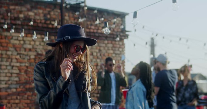 Close up of the Caucasian young charming woman in dark sunglasses and hat dancing and moving happily at the rooftop party at daytime. Outside.