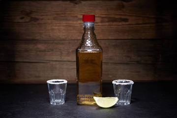 Gold tequila shots with lime fruits on wooden background. Bottle of tequila.