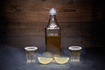Gold tequila shots with lime fruits on wooden background. Bottle of tequila. Smoke.