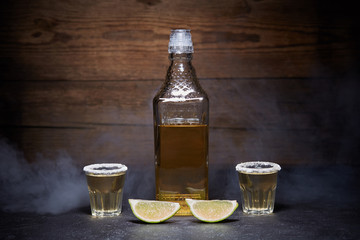 Gold tequila shots with lime fruits on wooden background. Bottle of tequila. Smoke.