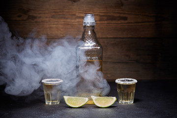 Gold tequila shots with lime fruits on wooden background. Bottle of tequila. Smoke.
