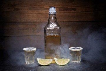 Gold tequila shots with lime fruits on wooden background. Bottle of tequila. Smoke.