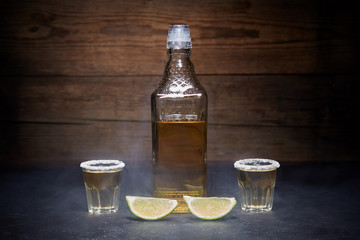 Gold tequila shots with lime fruits on wooden background. Bottle of tequila. Smoke.