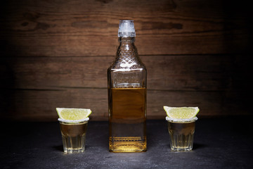 Gold tequila shots with lime fruits on wooden background. Bottle of tequila. Smoke.