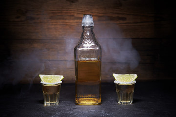 Gold tequila shots with lime fruits on wooden background. Bottle of tequila. Smoke.