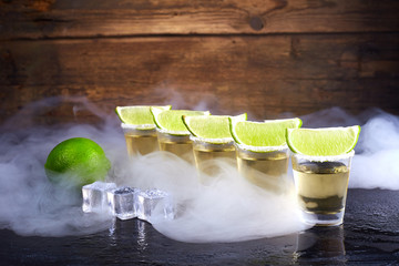 Mexican tequila gold in short glasses with salt, lime slices and ice on a wooden table. Smoke.
