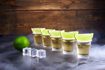 Mexican tequila gold in short glasses with salt, lime slices and ice on a wooden table. Smoke.
