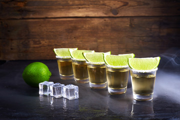 Mexican tequila gold in short glasses with salt, lime slices and ice on a wooden table. Smoke.