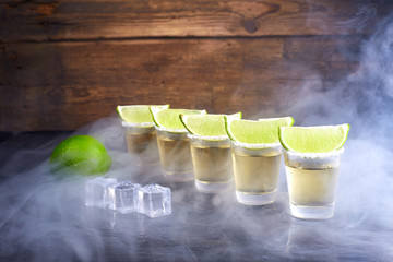 Mexican tequila gold in short glasses with salt, lime slices and ice on a wooden table. Smoke.