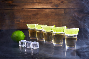 Mexican tequila gold in short glasses with salt, lime slices and ice on a wooden table. Smoke.