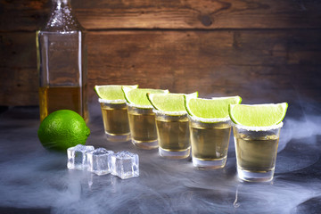 Mexican tequila gold in short glasses with salt, lime slices and ice on a wooden table. Smoke.