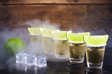 Mexican tequila gold in short glasses with salt, lime slices and ice on a wooden table. Smoke.