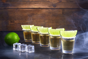 Mexican tequila gold in short glasses with salt, lime slices and ice on a wooden table. Smoke.