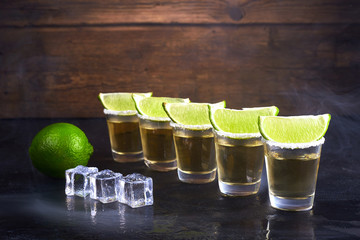Mexican tequila gold in short glasses with salt, lime slices and ice on a wooden table. Smoke.