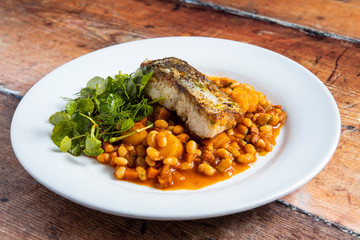 Hake with cannellini bean, chorizo and crab bisque stew with mini fondant potatoes on a white plate on a wooden floorboards background