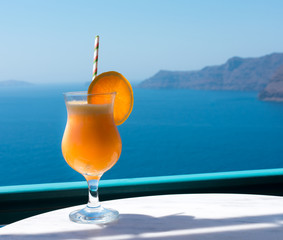 fresh orange juice just squeezed ready to be drunk. The orange juice is served on a table in a transparent glass cup with a straw and decorated with an orange slice. The Aegean sea at the background