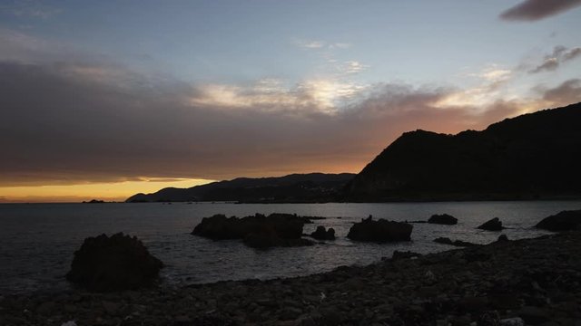 Red and orange sunrise timelapse video over the ocean, rocks, mountains in the city of New Zealand evening