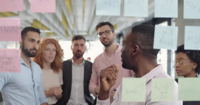 Male affrican team member counselling with colleagues and replacing sticky notes . Group of multi ethnic office workers standing and brainstorming at office meeting using glass board.