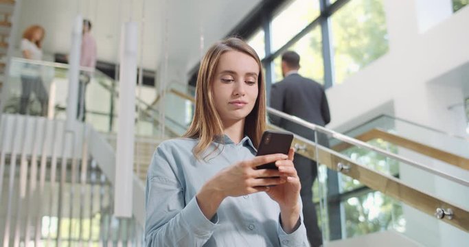 Young attractive female office worker using smartphone and standing near stairs. businesswoman typing messages at her mobile phone. Concept of people, work, technology.