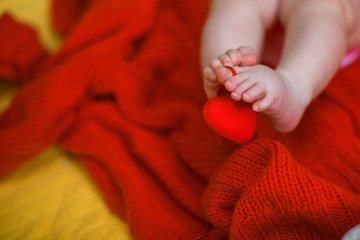 Red heart on baby legs. The legs of the newborn on a red background. A baby wrapped in a red blouse. Valentine's Day