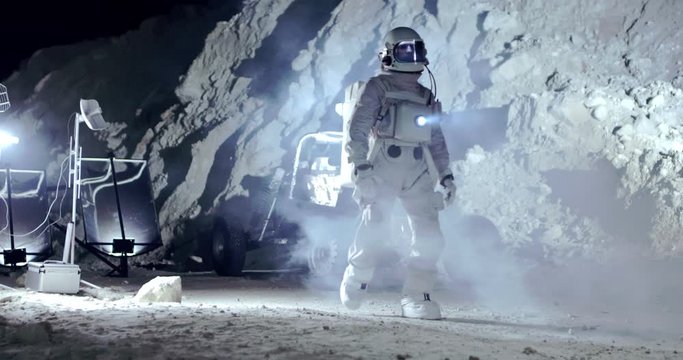Fully equipped NASA spaceman walking in the dark time in the Moon crater or valley of uninhabited planet at the station with equipment. Outdoors.