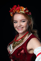 Naklejka premium portrait of attractive smiling woman in traditional costume in ukranian national wreath with flowers, red vest and golden necklace looking at camera isolated on black